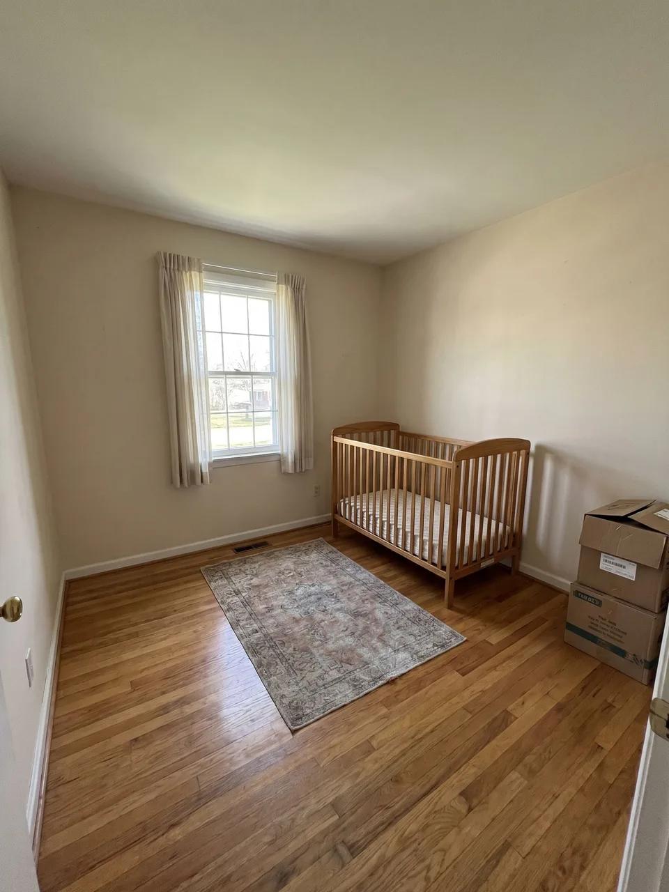 Small nursery with single window, thin curtains, small rug, wooden crib and moving boxes on hardwood floor