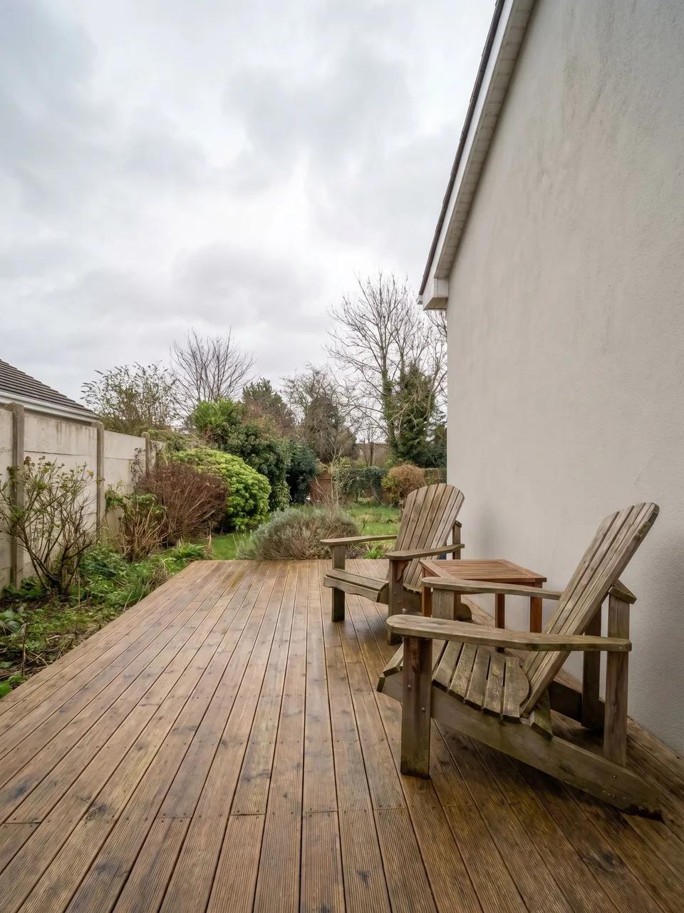 Long narrow wooden deck beside a pale exterior wall with two weathered wooden Adirondack chairs and garden beds along the left side.