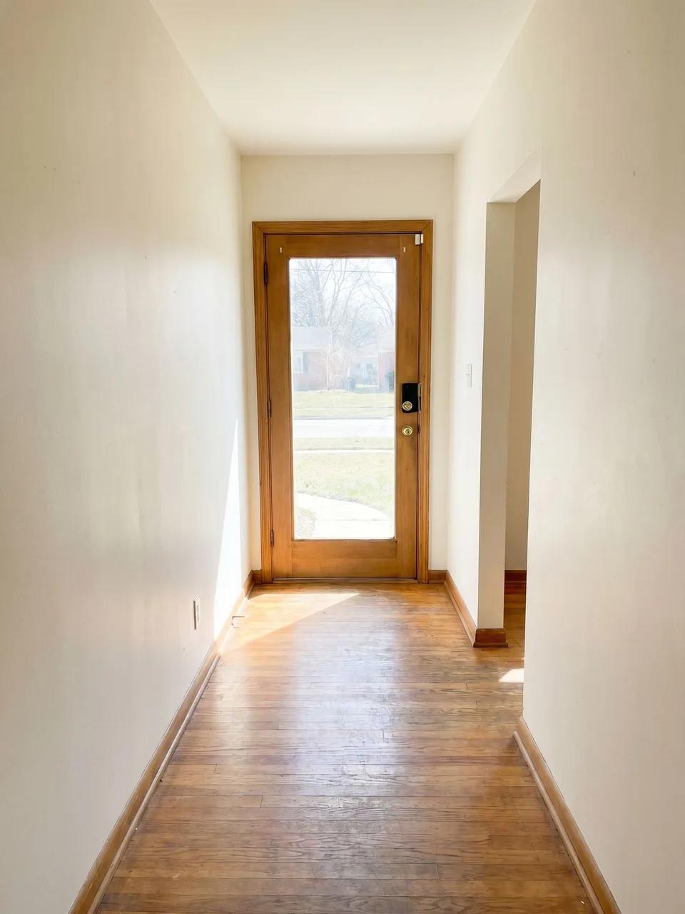 Long narrow entry with plain off-white walls, hardwood floor, and a full-glass wooden front door