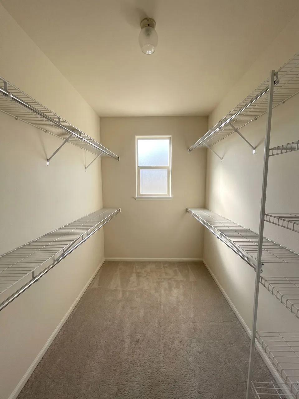 Narrow walk-in closet with white wire shelving on both sides, small centered window, beige carpet and single exposed ceiling bulb