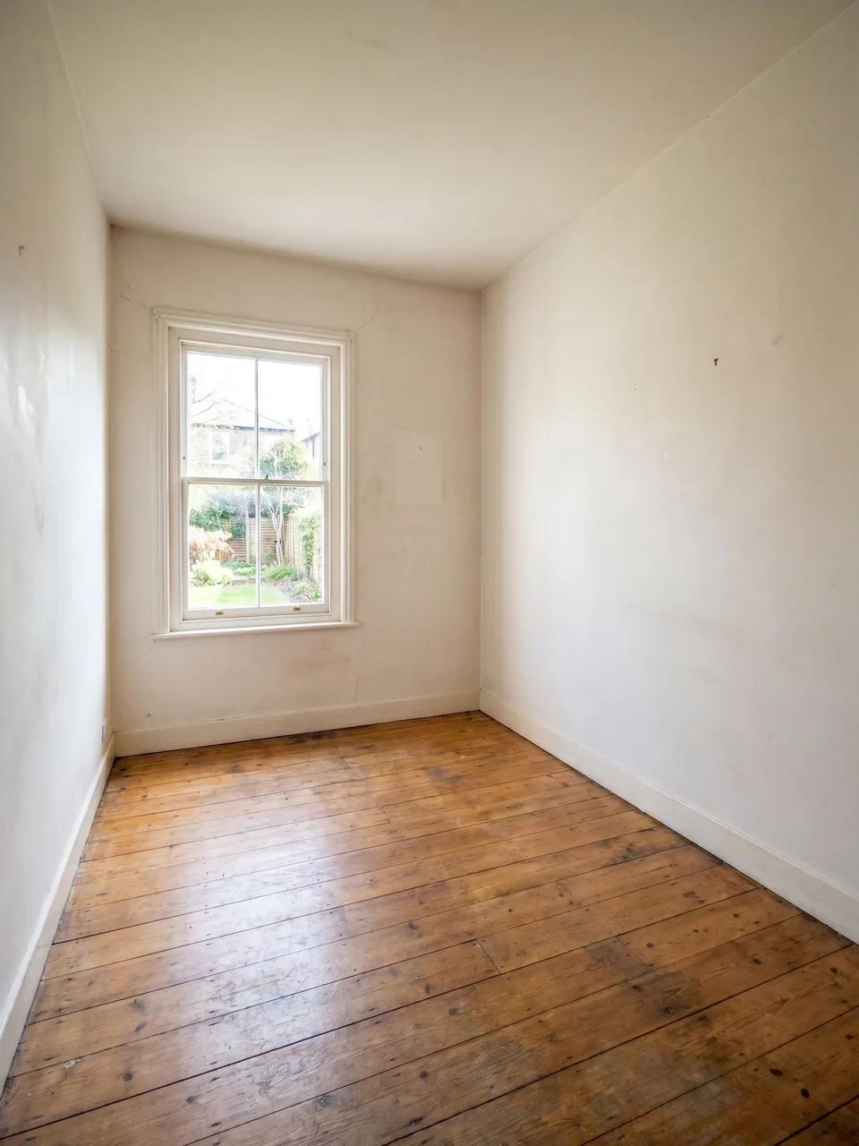 Empty narrow room with single sash window, white walls and worn wooden floorboards looking toward a garden.