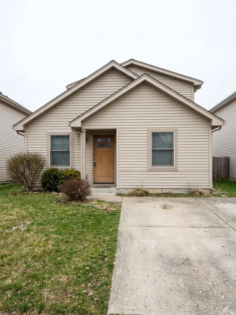 Small suburban front facade with beige vinyl siding, centered wooden door, two front windows, plain concrete driveway and sparse shrubs.