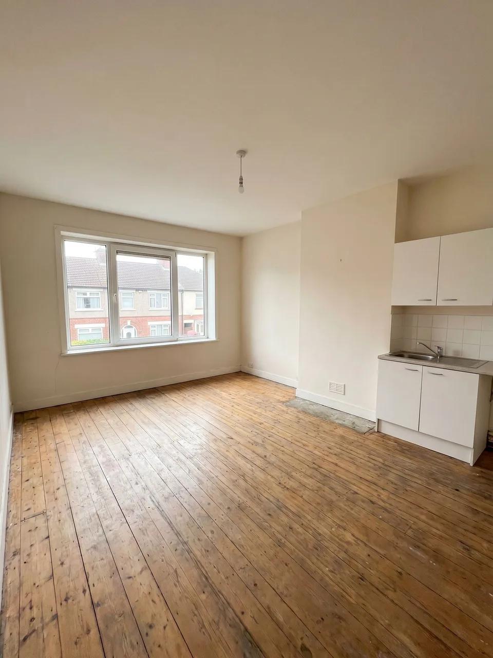 Empty studio with natural plank floor, large front window and small white kitchenette on the right