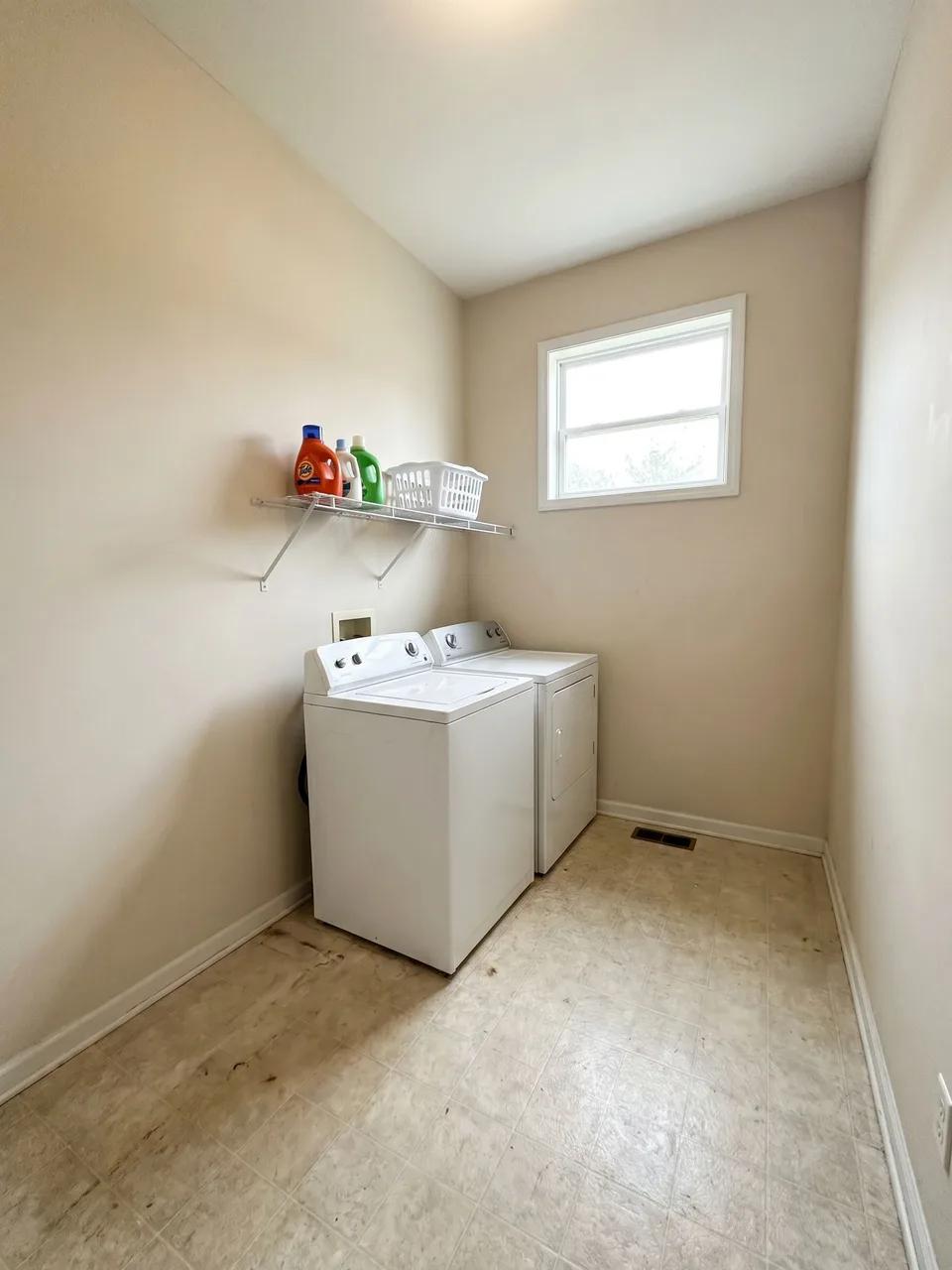 Narrow laundry with white washer and dryer under a single wire shelf and worn vinyl flooring, small window on far wall.