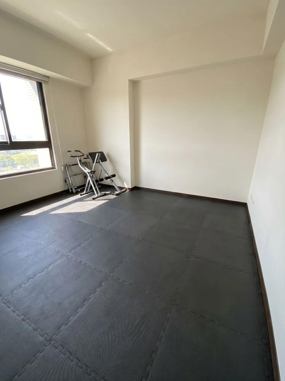 Empty home gym with interlocking black floor tiles, a window on the left and a small folding exercise device near the corner