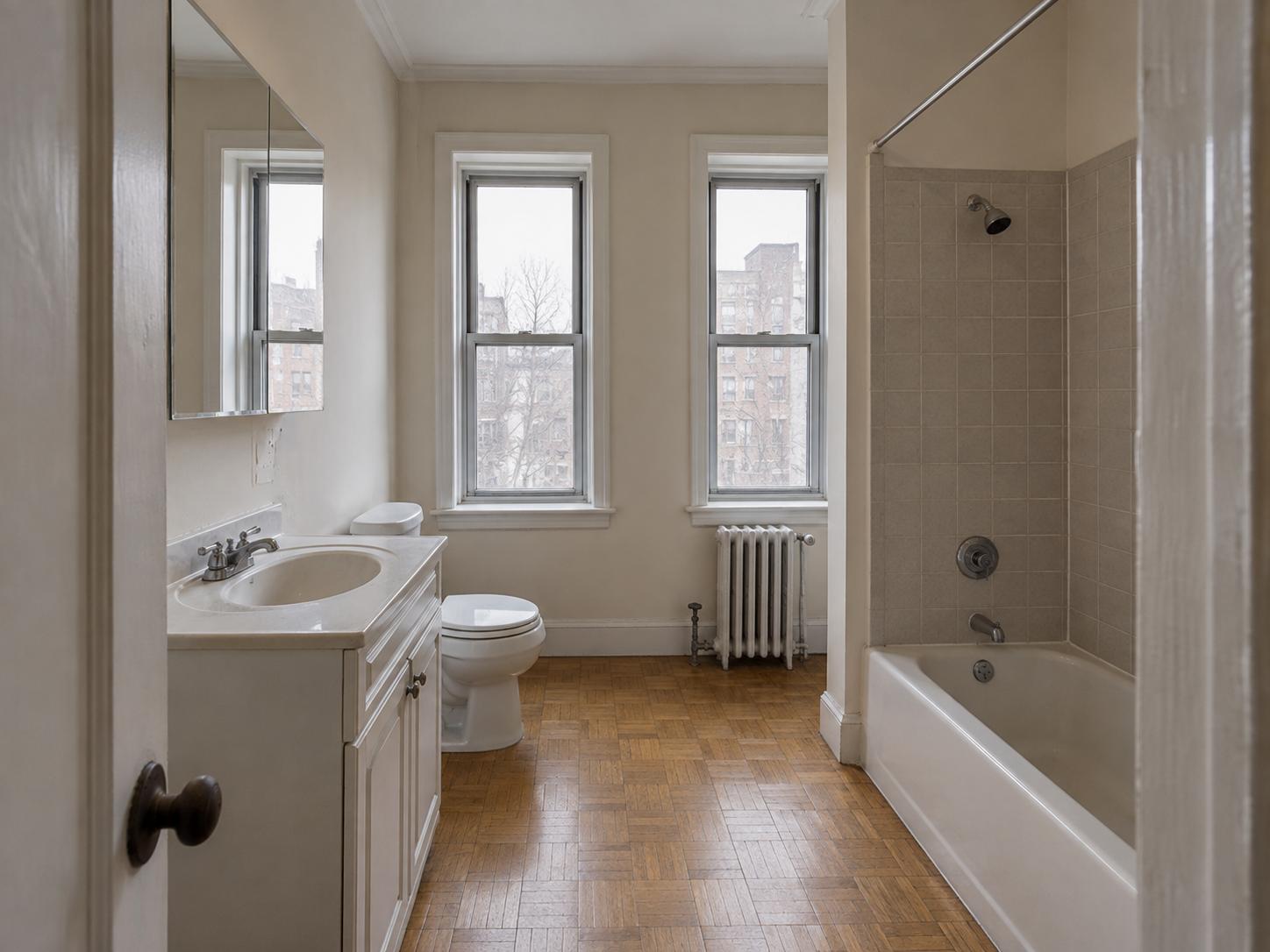 Older urban bathroom with parquet floor, built-in white vanity, mirrored medicine cabinet, two tall windows and tiled tub surround