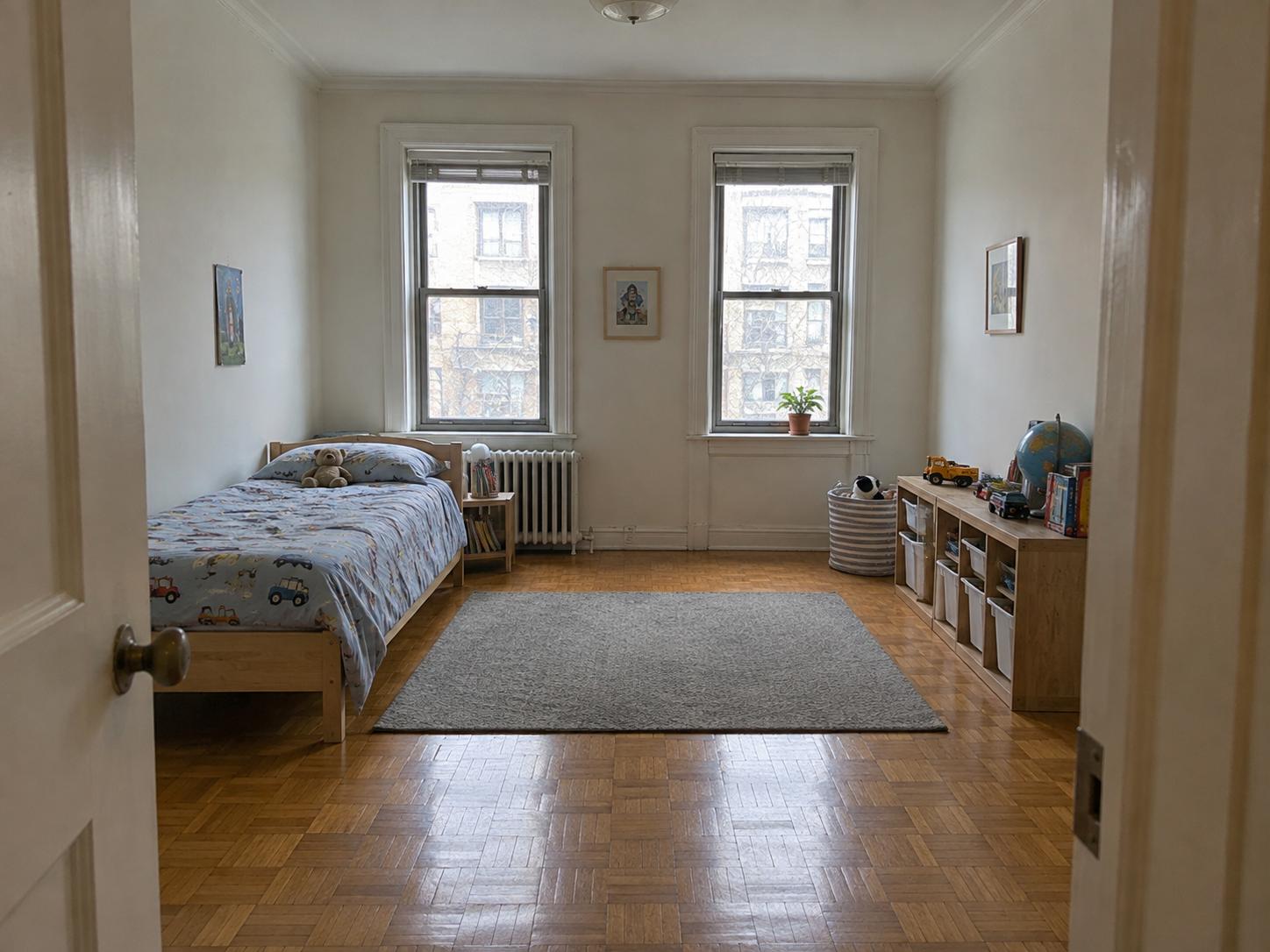 Simple kids room with vehicle-print duvet, low wooden bed, grey rug, radiator under two windows and toy cubby on the right