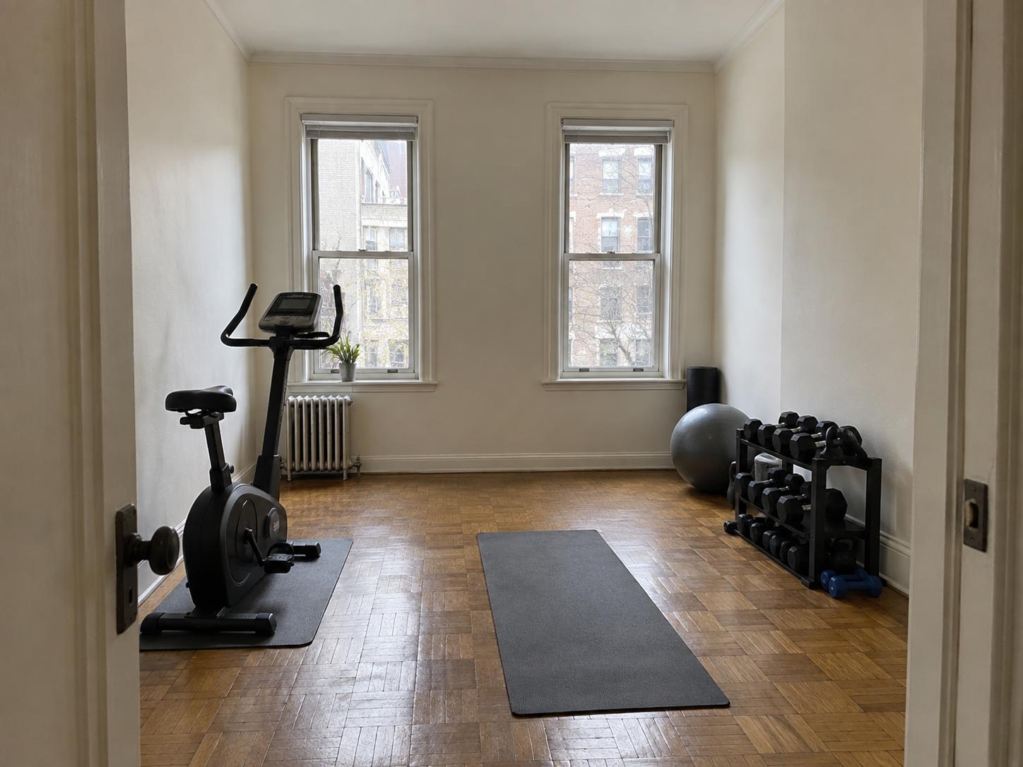 Small home gym with parquet floor, upright bike left, yoga mat center, dumbbell rack and exercise ball right, two sash windows above radiator.