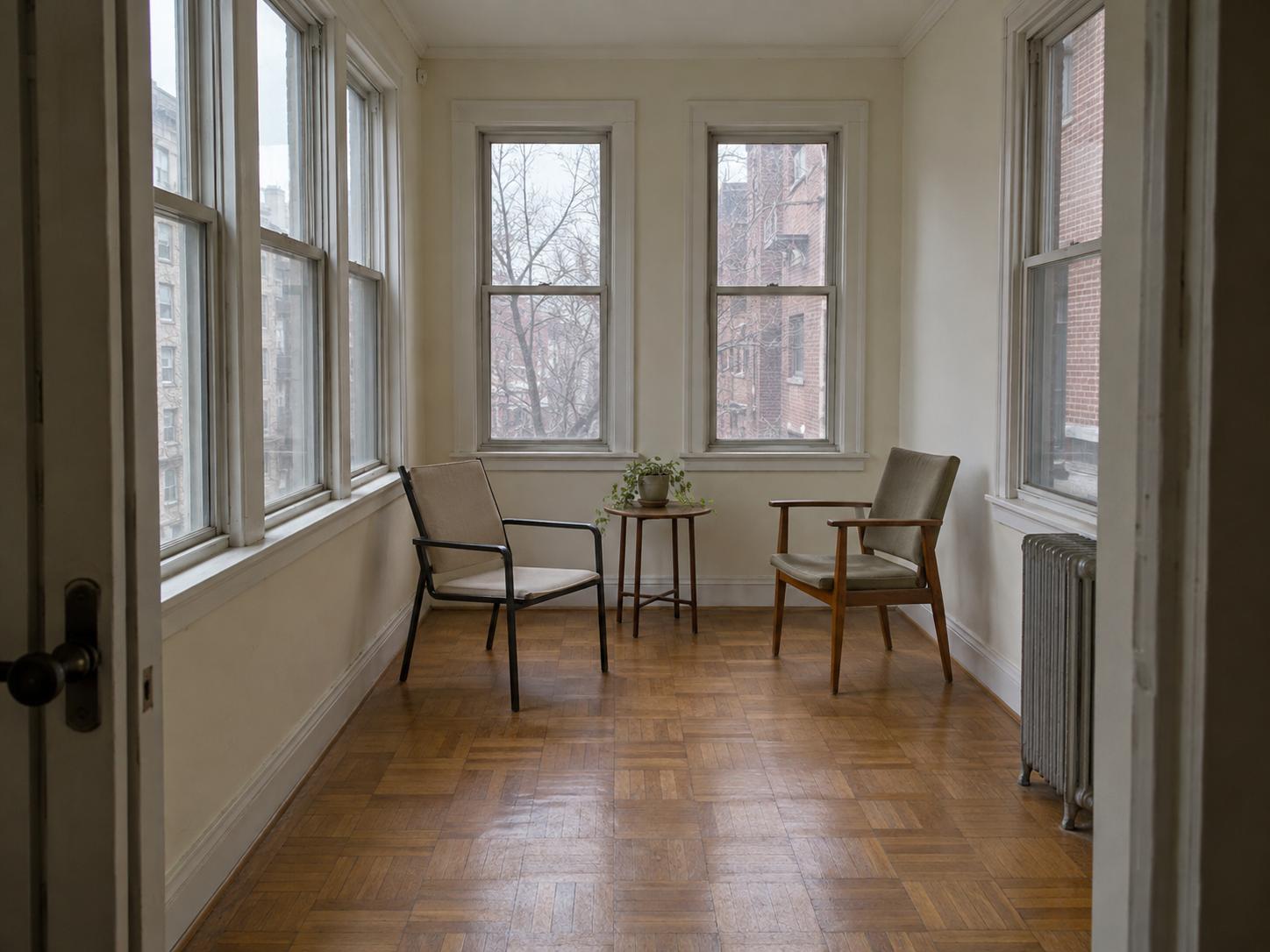 Narrow sunroom with three tall windows, parquet floor, two chairs and small round table, radiator at right