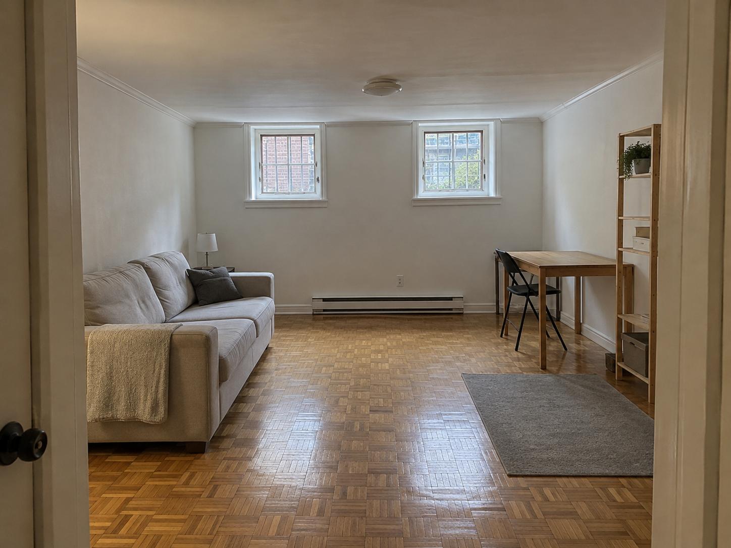 Long narrow basement with two small high windows, beige sofa left, desk right, parquet floor and minimal furnishings