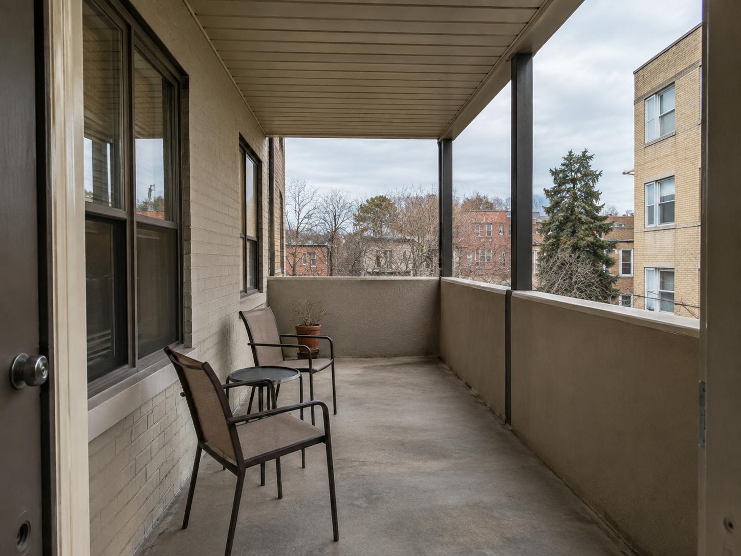 Narrow covered patio with two metal-frame chairs, small round table, concrete floor and beige brick wall