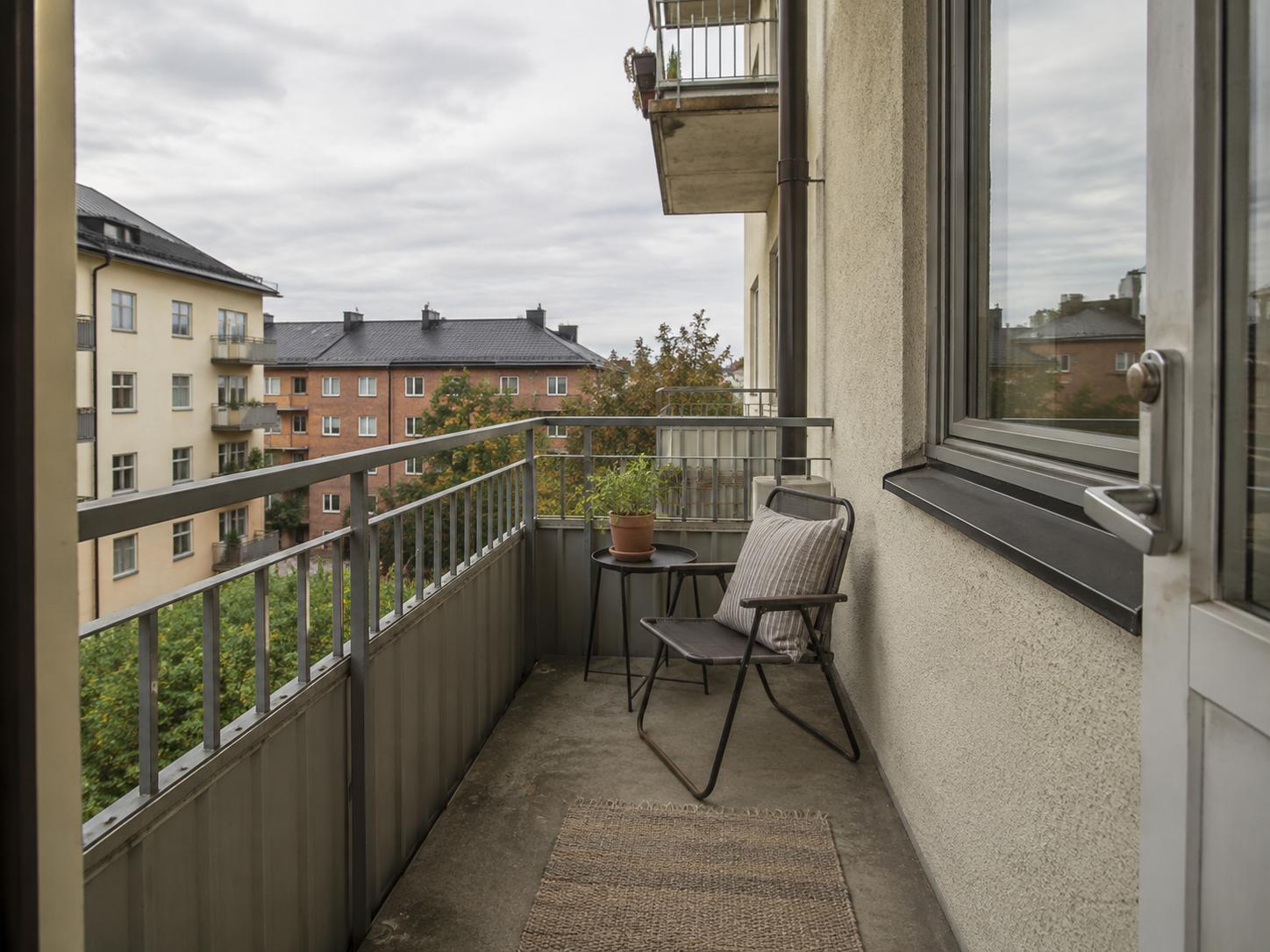 Narrow apartment balcony with single black folding chair, small round black table, jute rug, metal railing and concrete floor