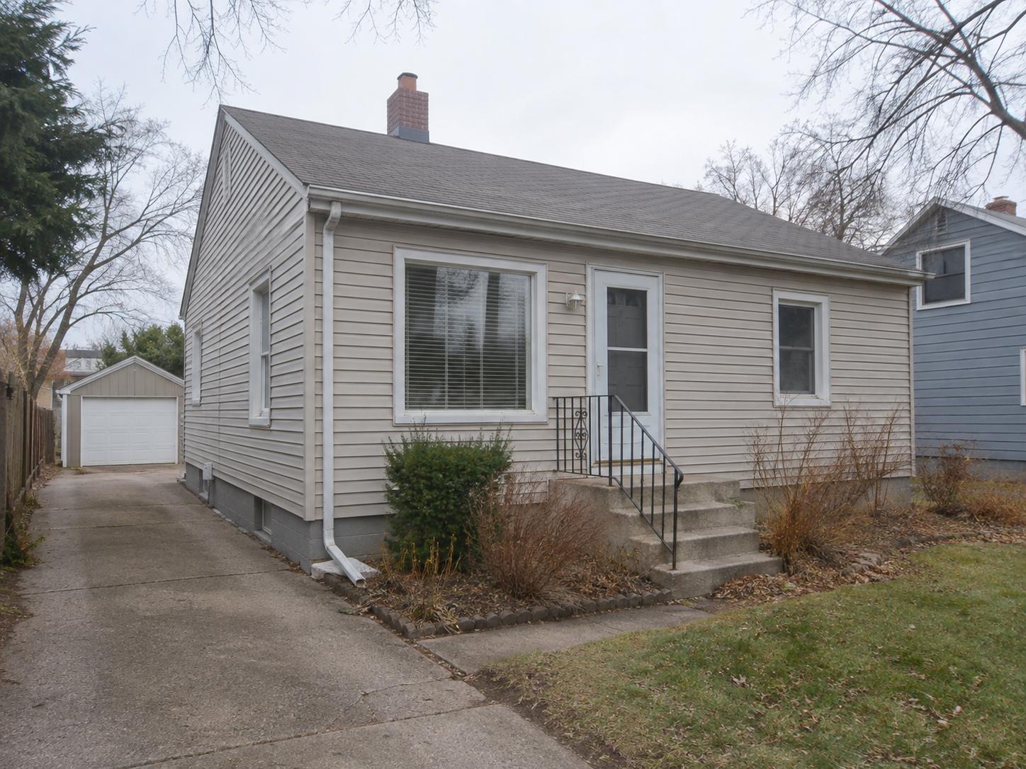 Single-story suburban house exterior with concrete steps, iron handrail, beige siding and narrow shrub planting bed by the driveway.
