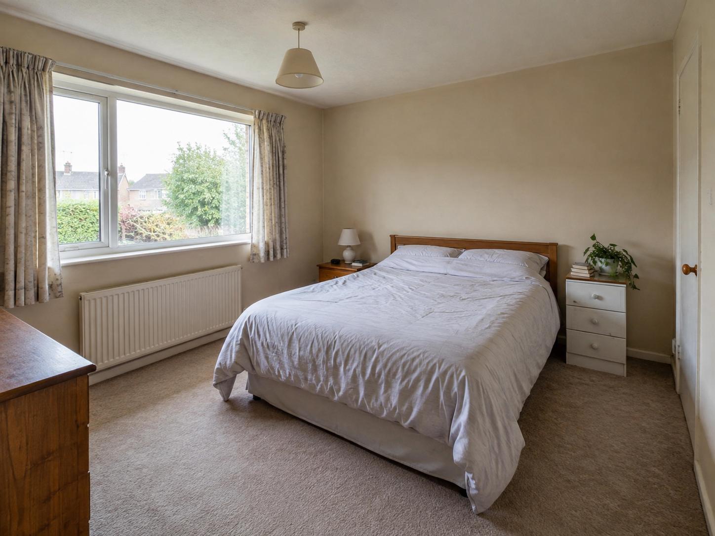 Compact bedroom with beige walls, simple timber headboard, white bedding, small bedside tables and a large window with radiator below