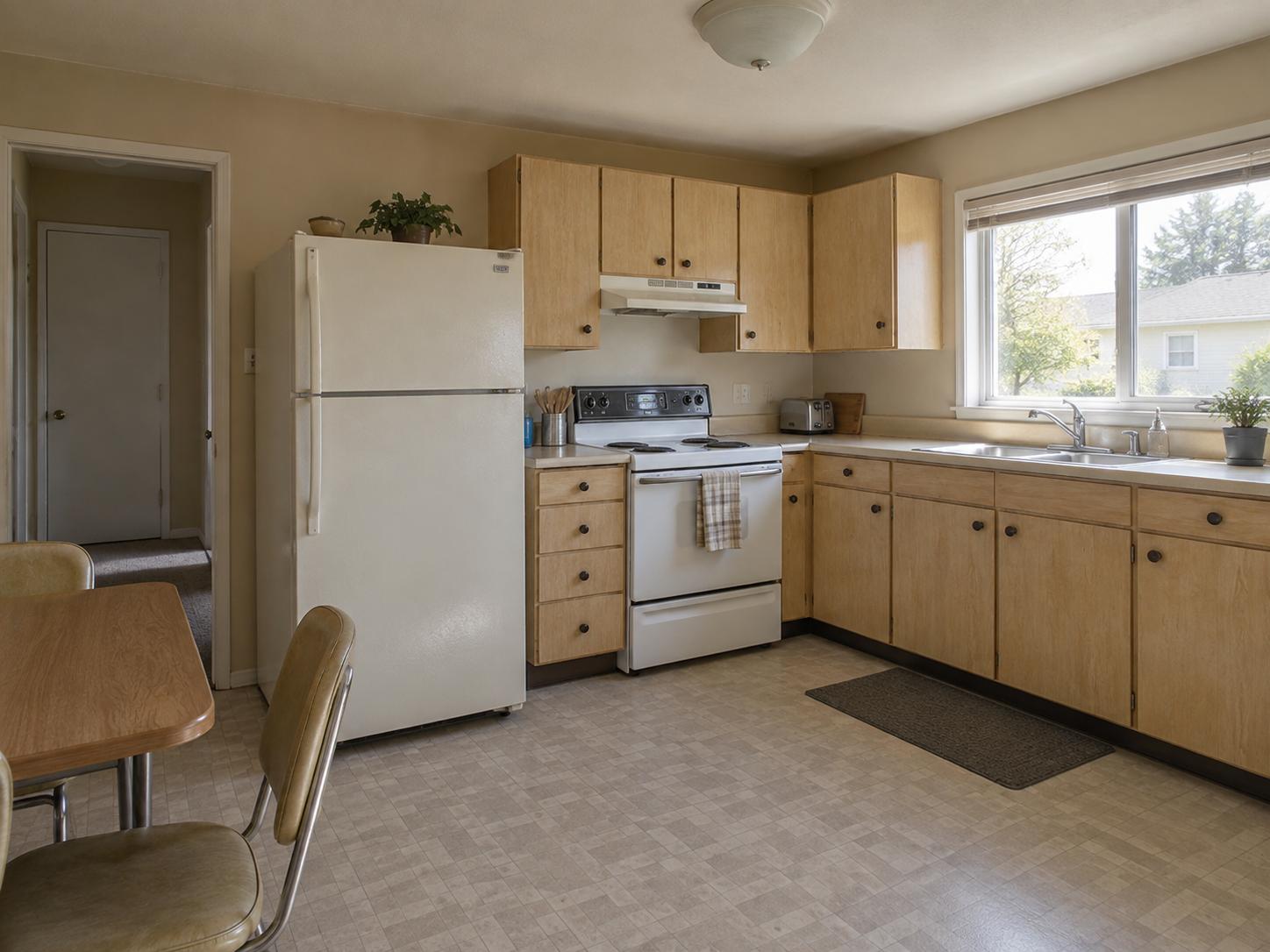 Compact mid-century kitchen with light maple cabinets, white fridge and range, vinyl floor and large sink window