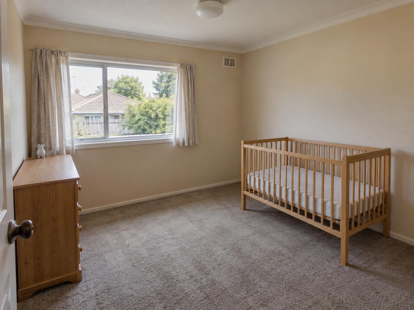 Compact nursery with beige walls, wooden crib on right, small dresser by window, beige carpet and wide window with light curtains.