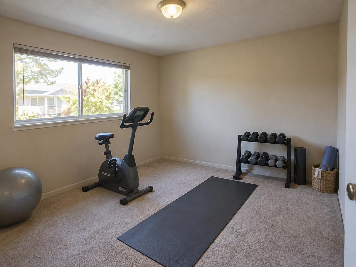 Small carpeted home gym with exercise bike, yoga mat, dumbbell rack and exercise ball under a wide window