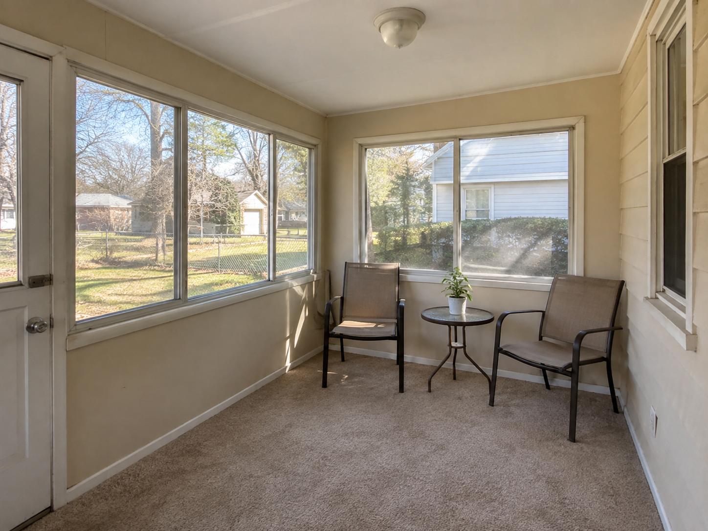 Narrow sunroom with beige walls, carpet floor, two metal-framed chairs and a small round table by large windows.