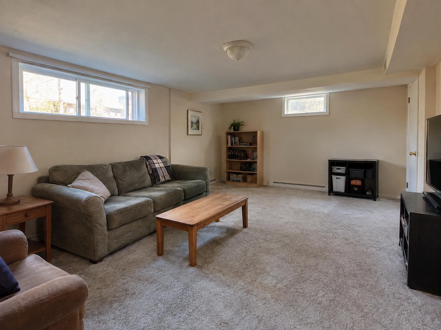 Basement living room with olive-green sofa, wooden side table, low horizontal windows and beige carpeting.