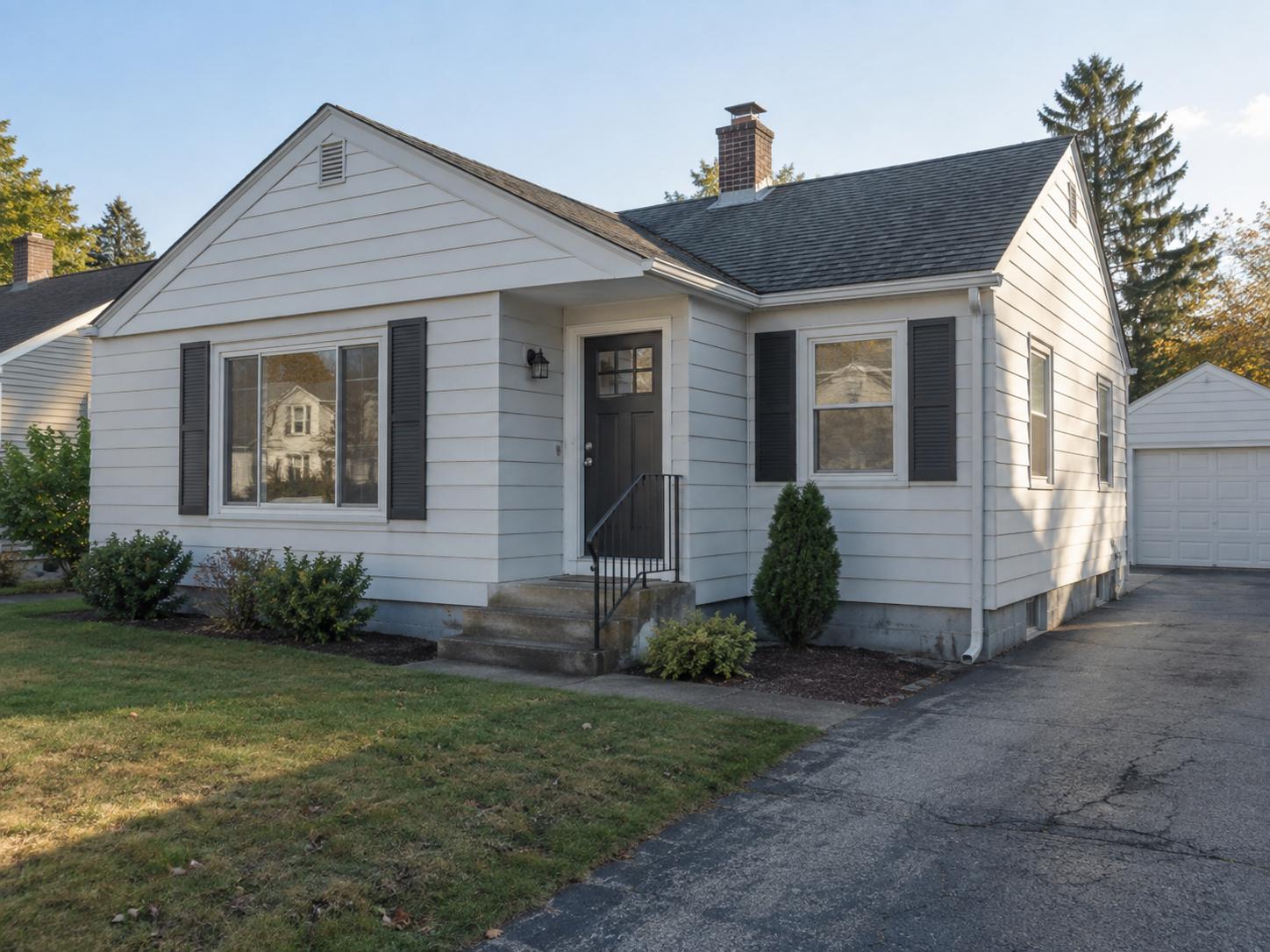 Small single-story white siding cottage with black shutters, plain black front door, concrete steps and sparse foundation plantings.
