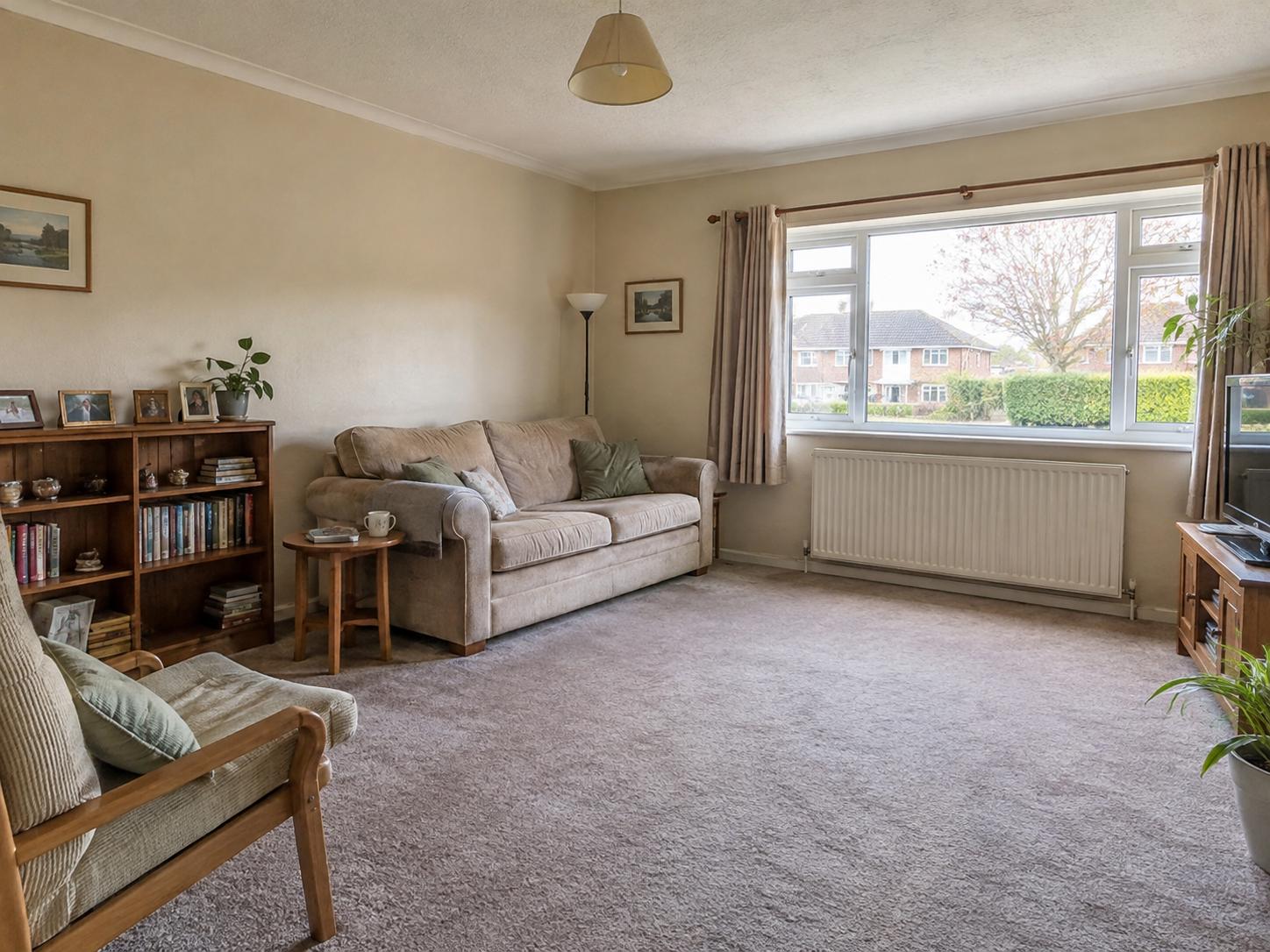 Compact family sitting room with beige three-seat sofa, wooden bookcase, single armchair and a large front window
