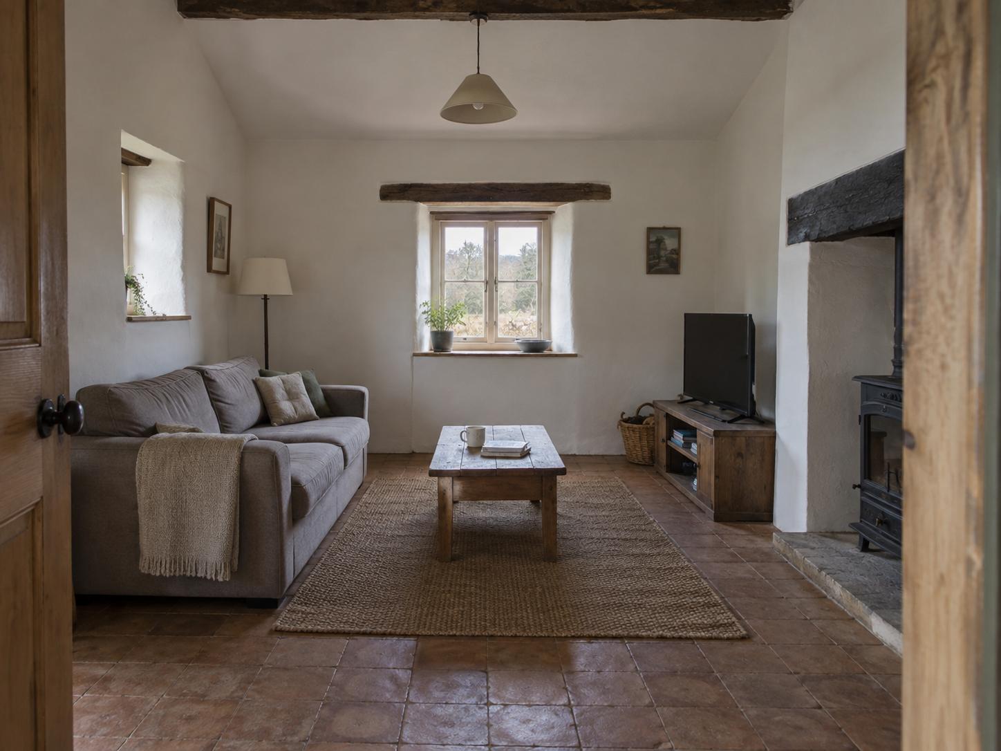Small rural cottage living room with gray sofa, jute rug, wooden coffee table, dark TV cabinet and visible ceiling beam.