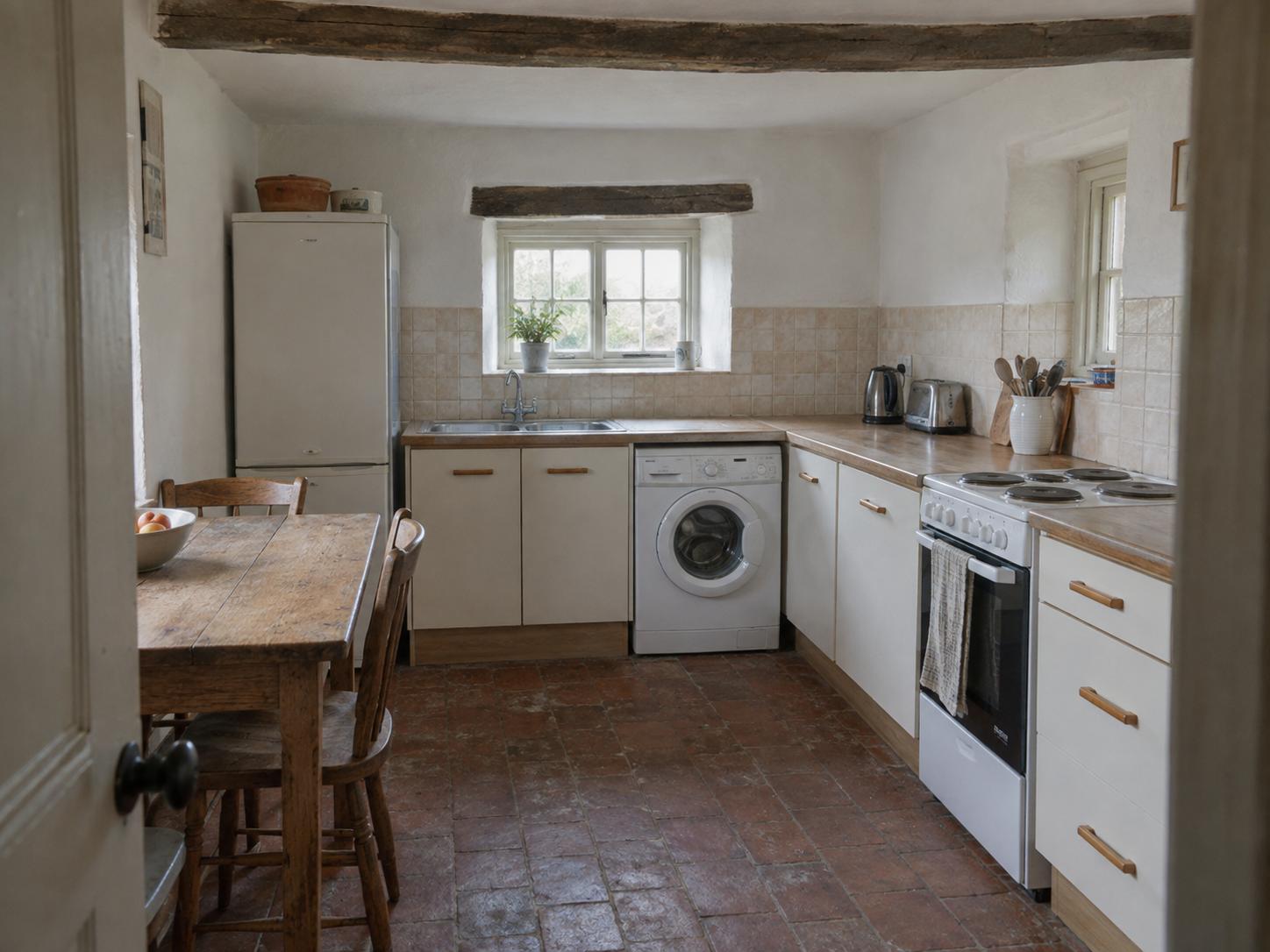 Small cottage kitchen with flat-front cream cabinets, wooden table and chairs, washing machine under counter, terracotta tile floor and exposed ceiling beams.