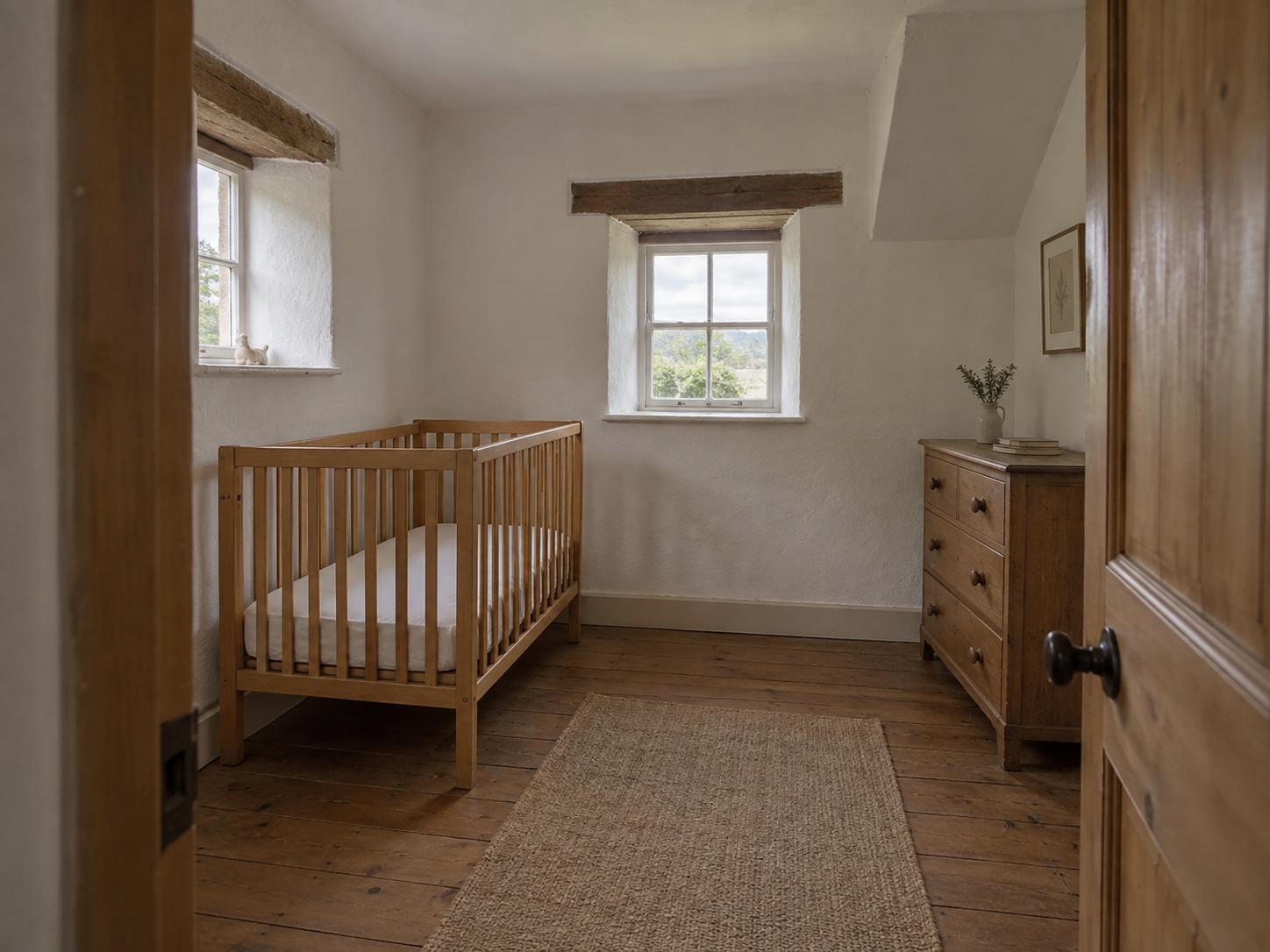 Compact cottage nursery with pale wood crib, dark woven runner, small central mullioned window and darker wooden dresser under sloped ceiling.