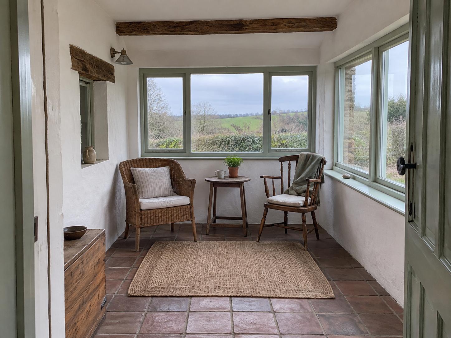 Small rural sunroom with two chairs, round table, jute rug, exposed beam and pale green window frames looking out to fields