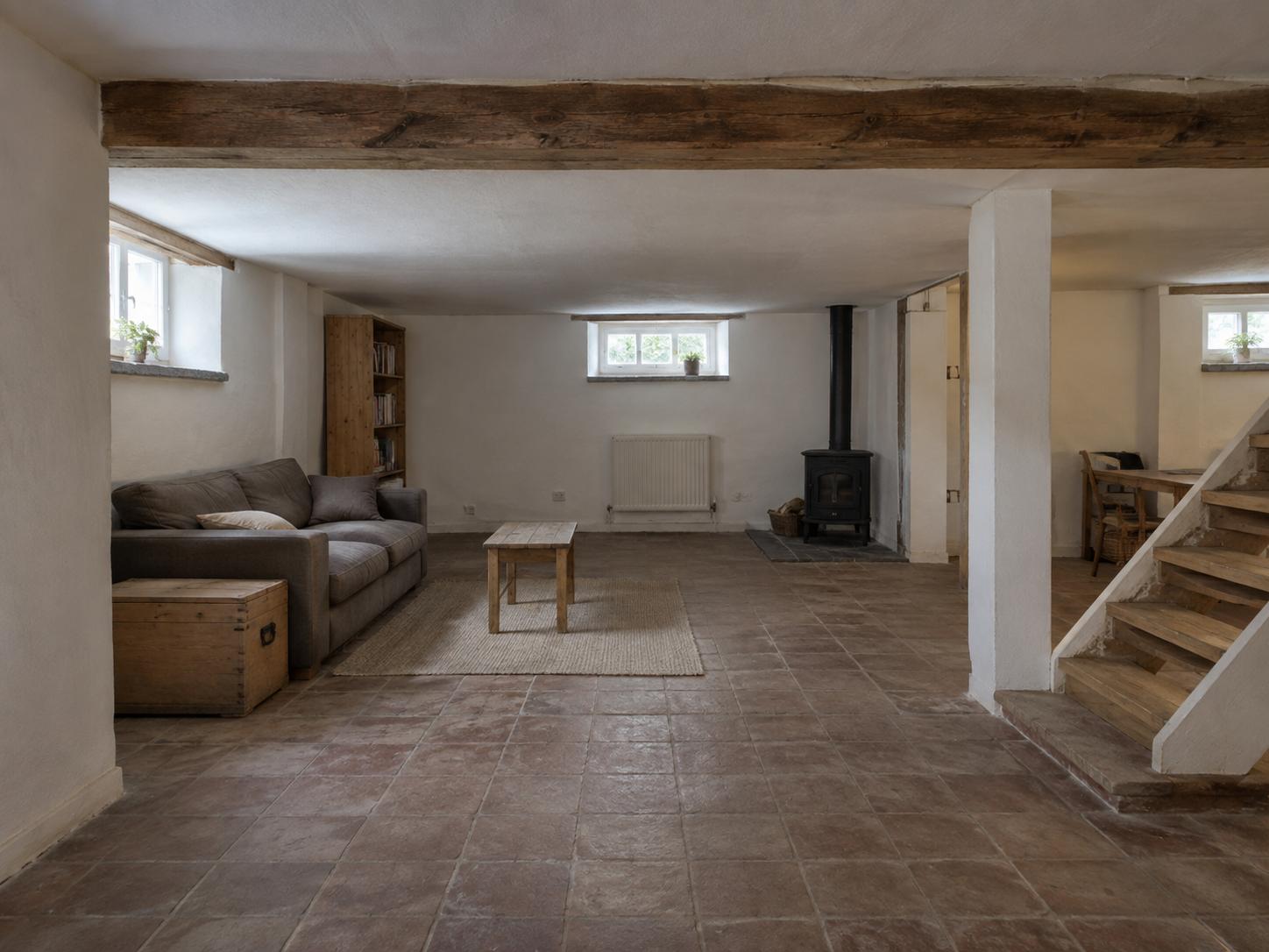 Low-beamed basement with grey sofa, trunk side table, bookcase, wood-burning stove and tiled floor