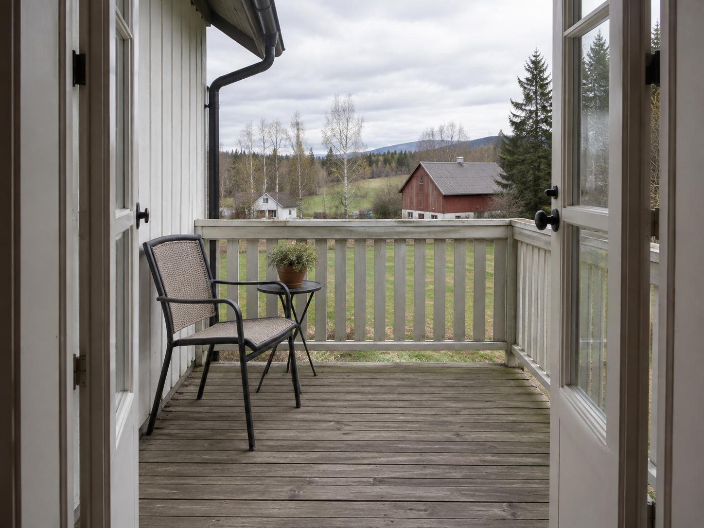 Narrow wooden balcony with a single metal chair, small round side table, slatted railing and view of fields and red barn.