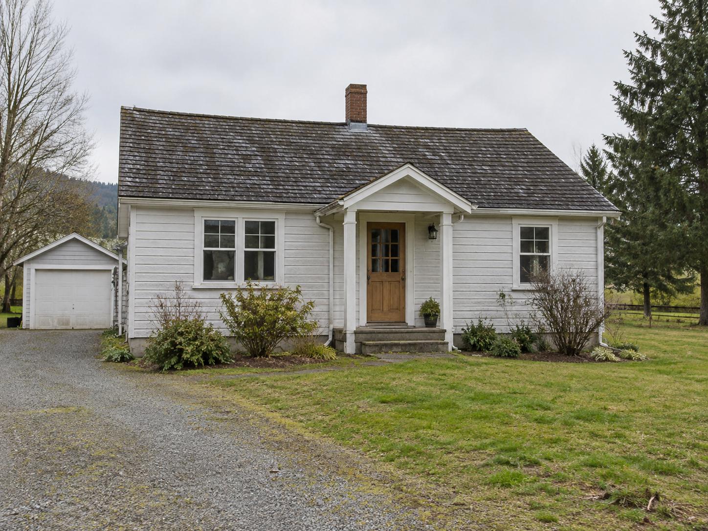 White rural cottage exterior with centered gabled porch, plain wood door, concrete steps, sparse shrubs and gravel driveway to a small garage.