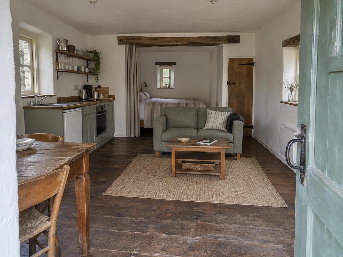 Small cottage studio entry showing dark-stained kitchenette, green sofa, wooden coffee table and jute rug under exposed beam.
