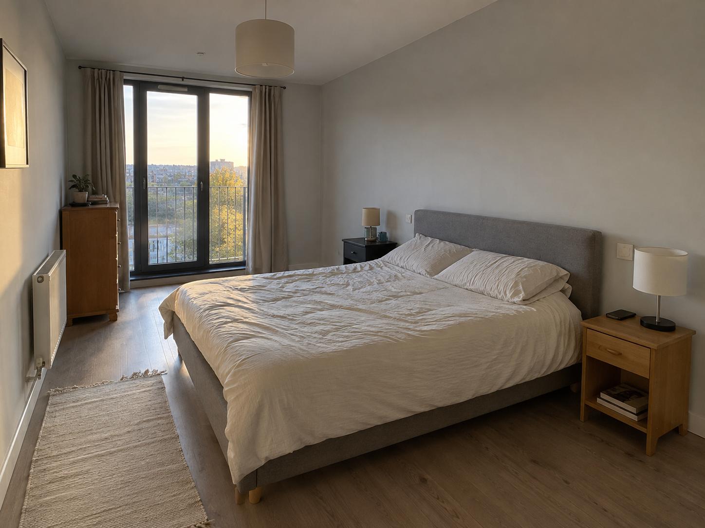 Neutral bedroom with grey upholstered bed, mismatched wooden bedside tables, narrow dresser by balcony doors and a runner rug along the bed side.