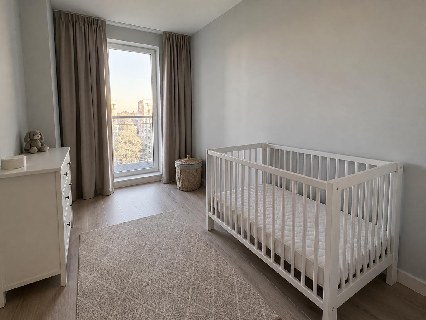 Small nursery with white crib, white dresser, plain beige rug and tall window with taupe curtains