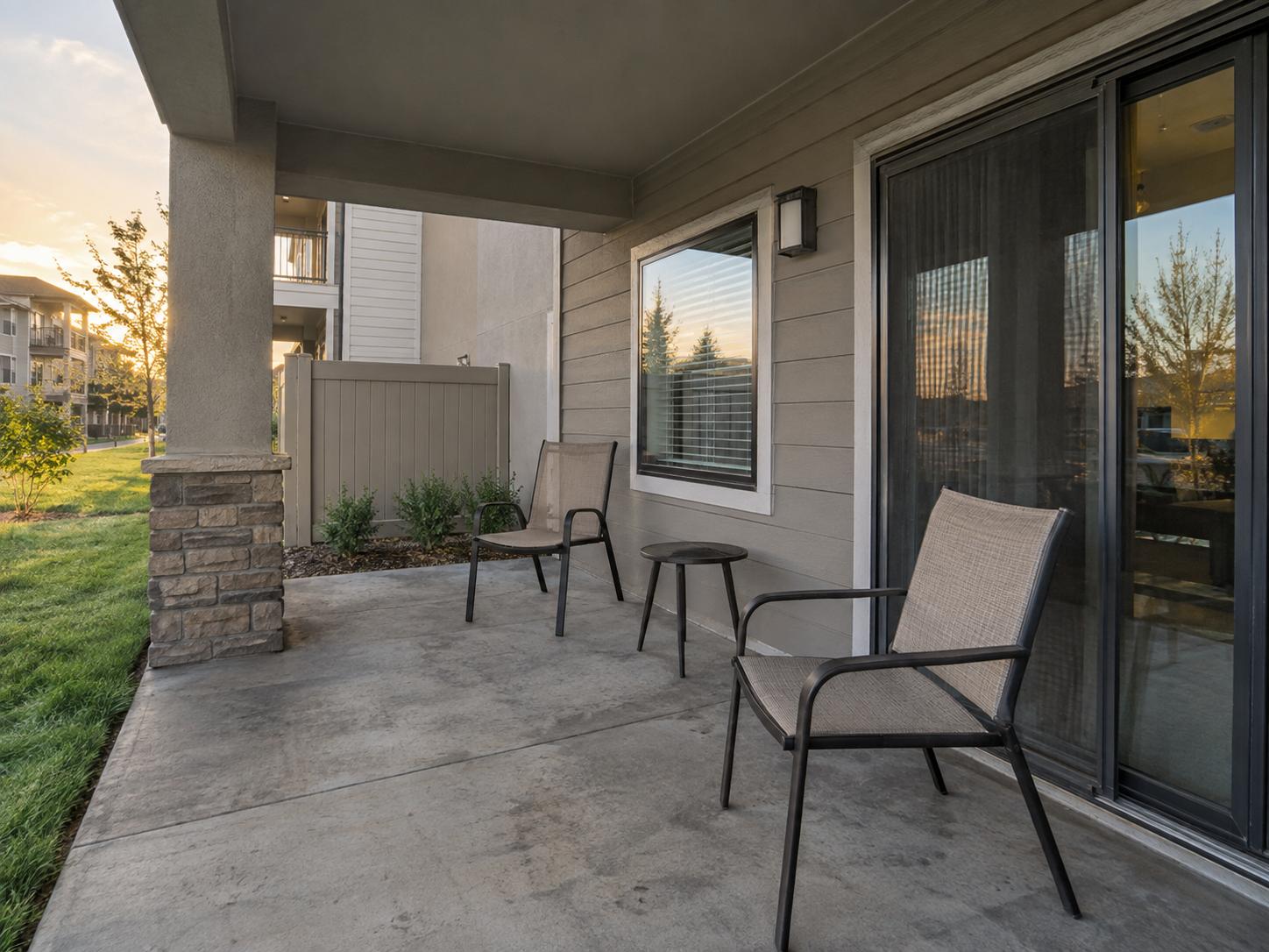 Covered apartment patio with stone column, two metal-framed chairs, small round table, concrete floor and sliding glass door