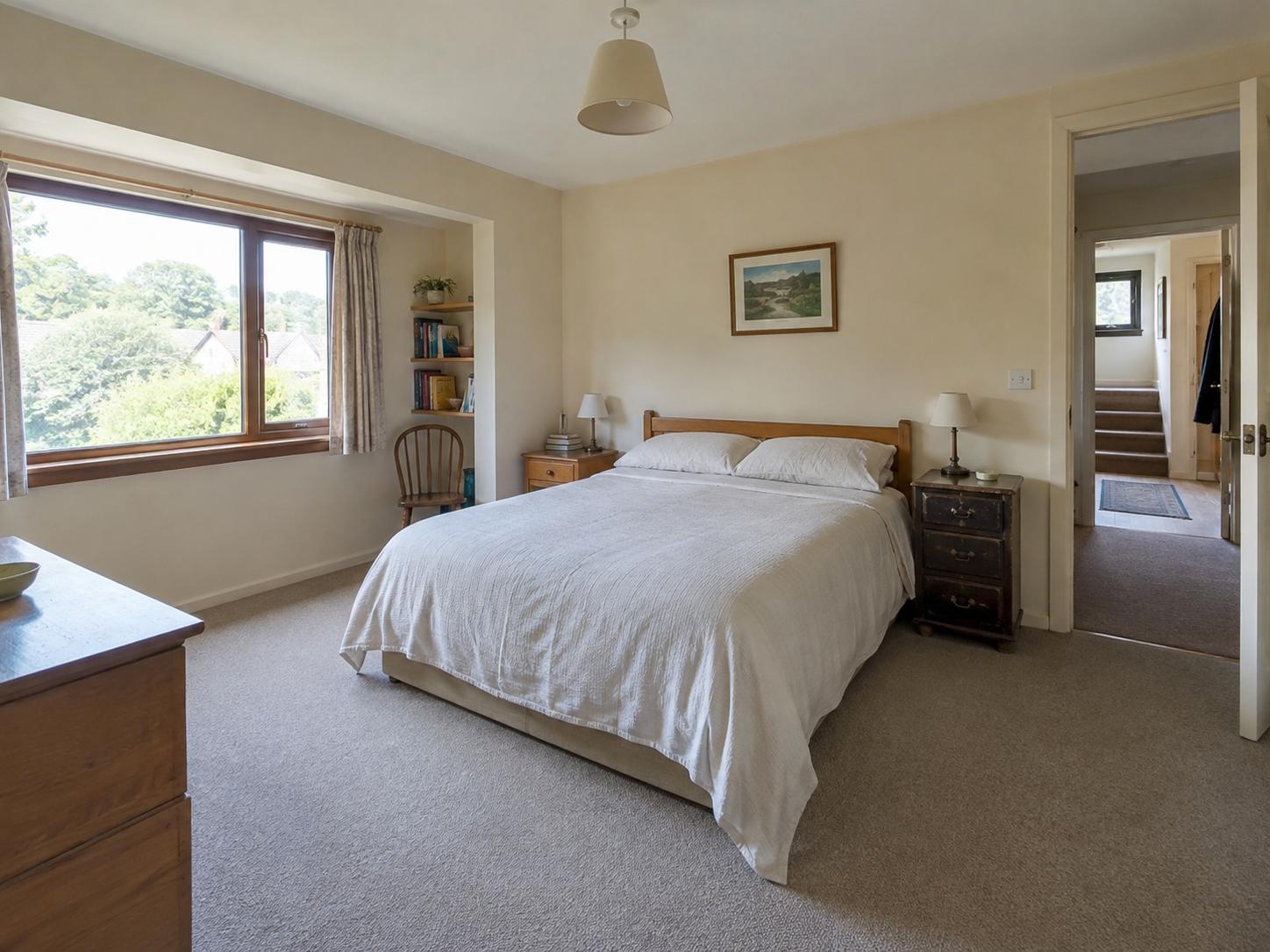 Bedroom with wooden bedframe, two bedside tables, window alcove with chair and bookshelves, pendant light