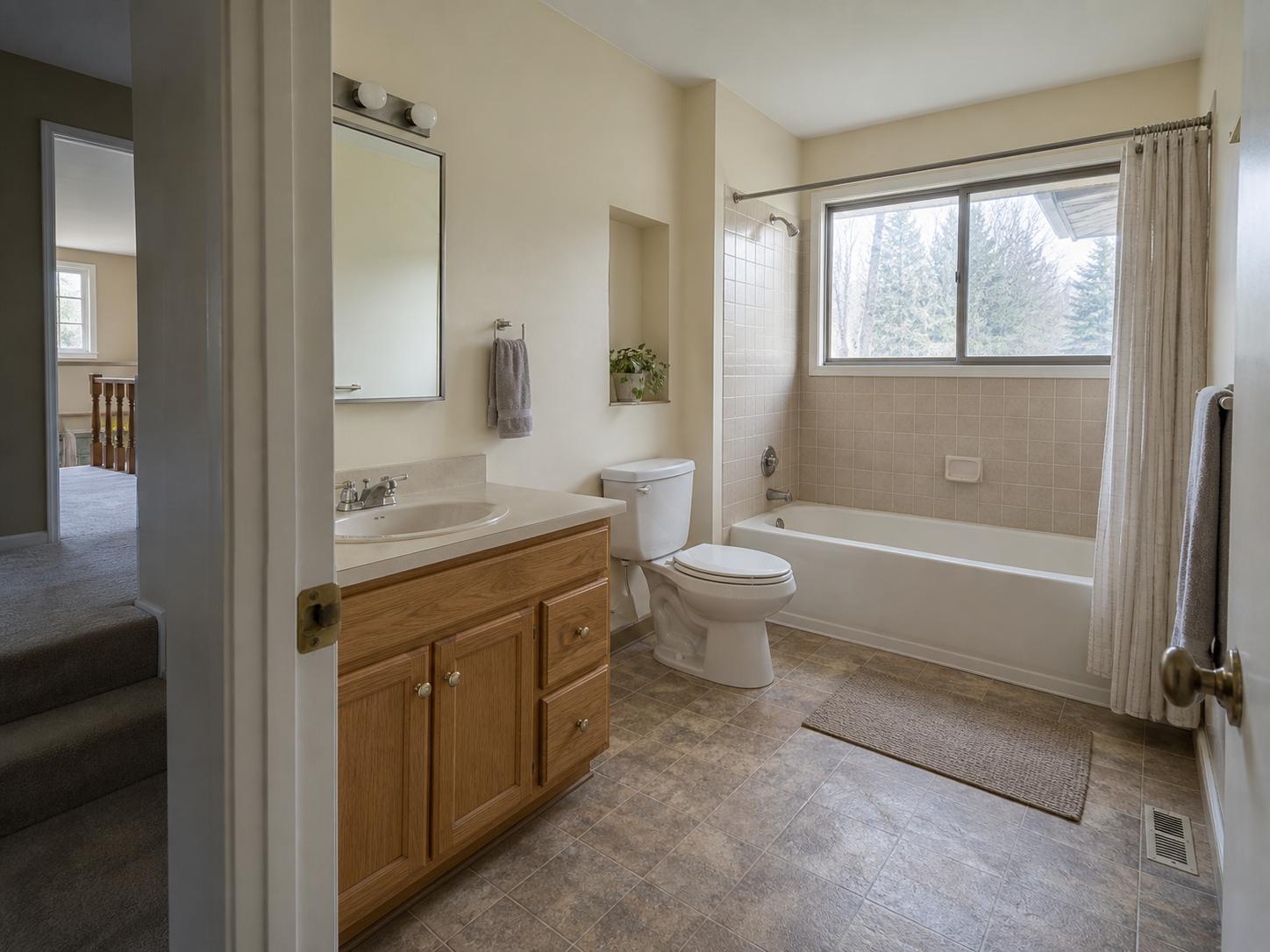 Modest split-level bathroom with oak base vanity, framed mirror, tub with short beige square tile surround and single window.