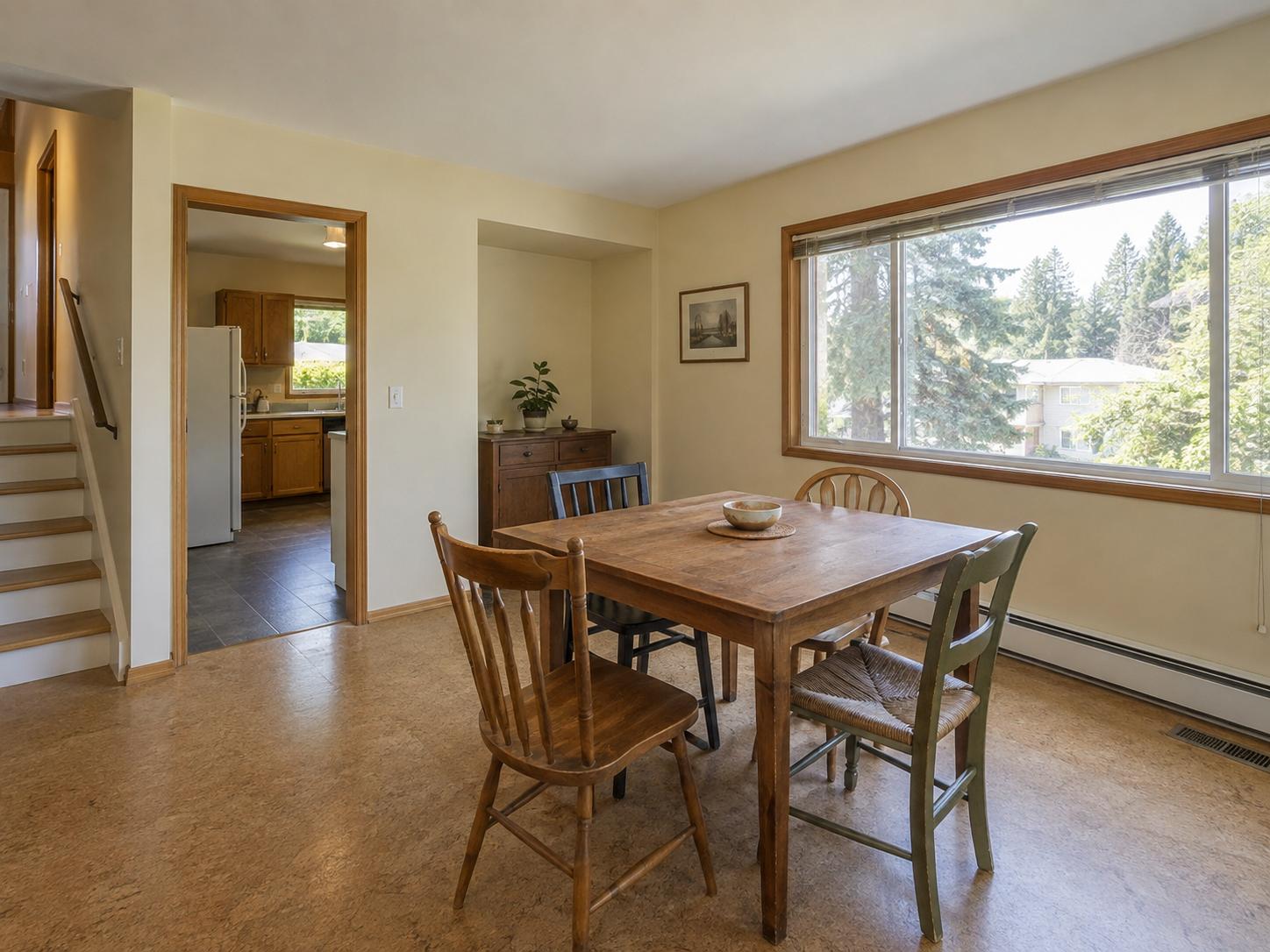 Modest split-level dining area with large window, plain walls, wooden table and four mixed wood chairs