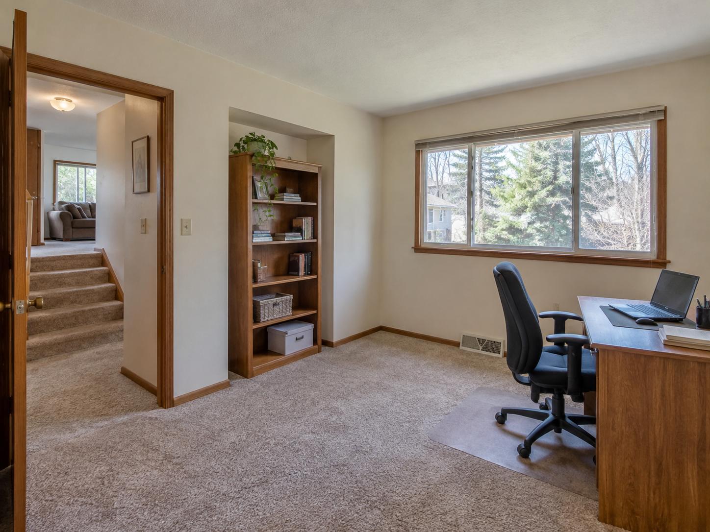 Modest home office with beige carpet, dark wood bookcase in recessed niche and a dark desk by a large window.