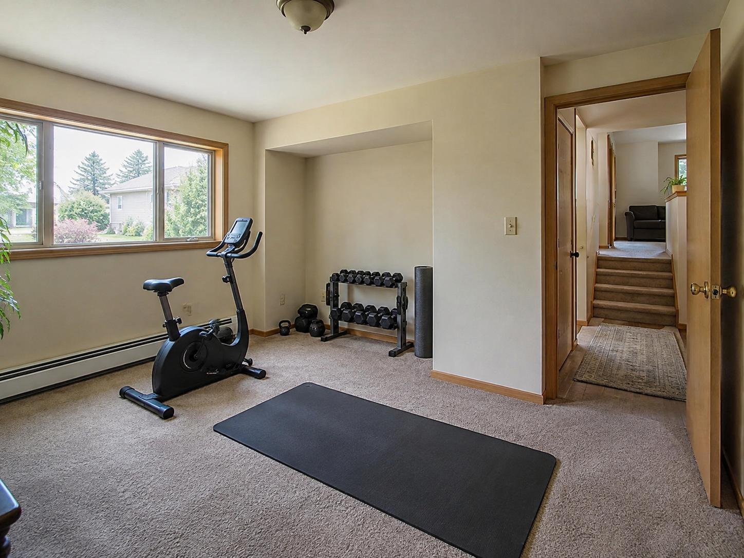 Small home-gym with exercise bike by a large window, single black mat on carpet, and free weights stored on a rack in a recessed alcove.