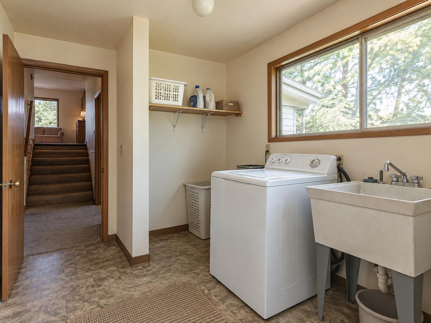 Modest laundry room with top-loading washer, freestanding utility sink, single open shelf, window with wood trim and vinyl flooring.