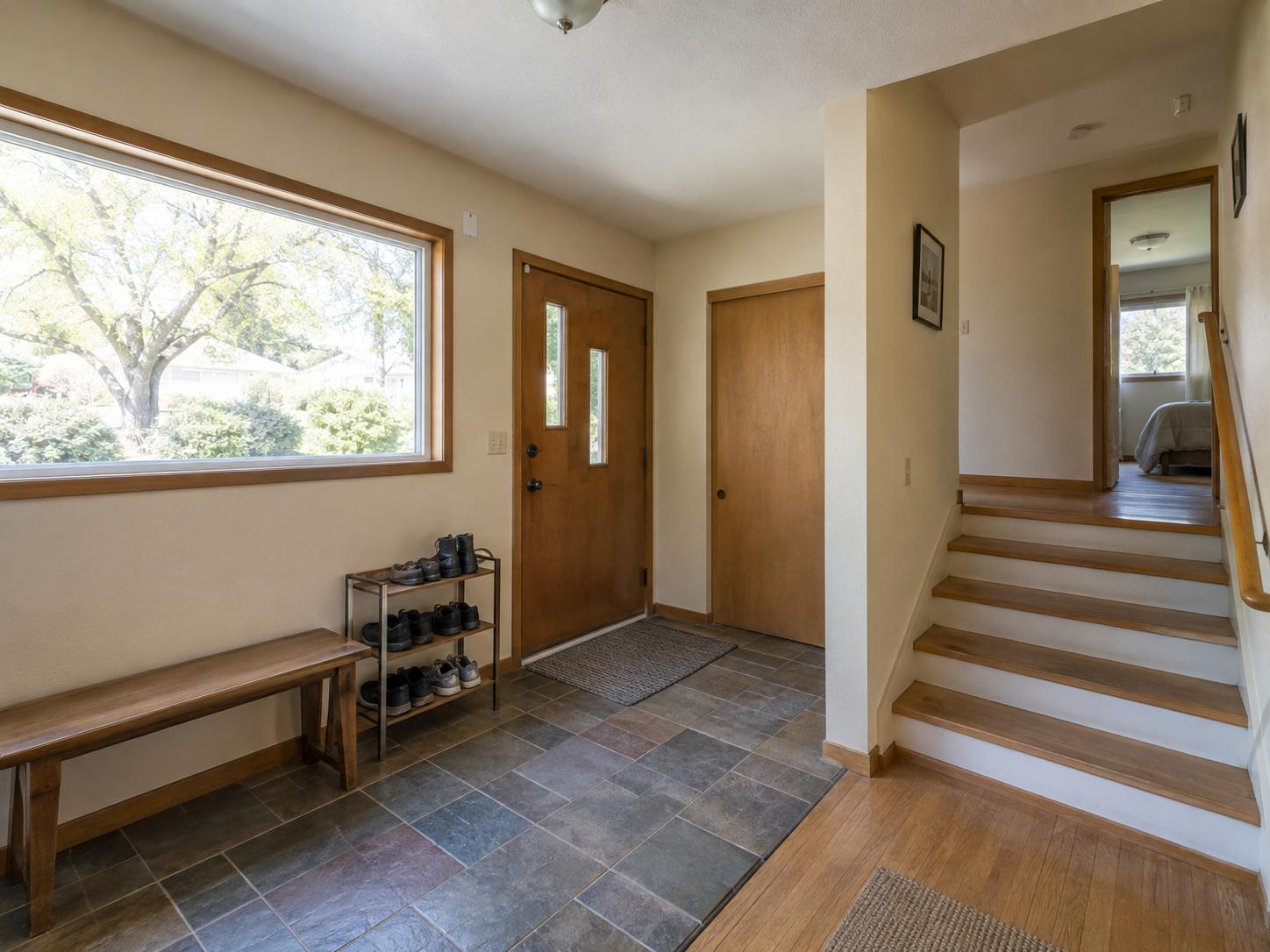 Modest split-level entryway with large picture window, wooden front door, metal shoe rack and simple wooden bench on slate tile