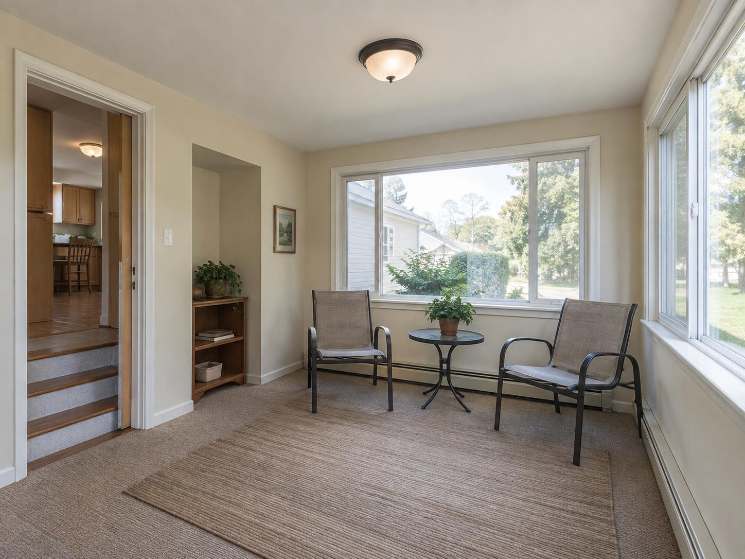 Small sunroom with two mesh chairs, round metal table, low bookshelf in a recessed niche and large front and side windows.