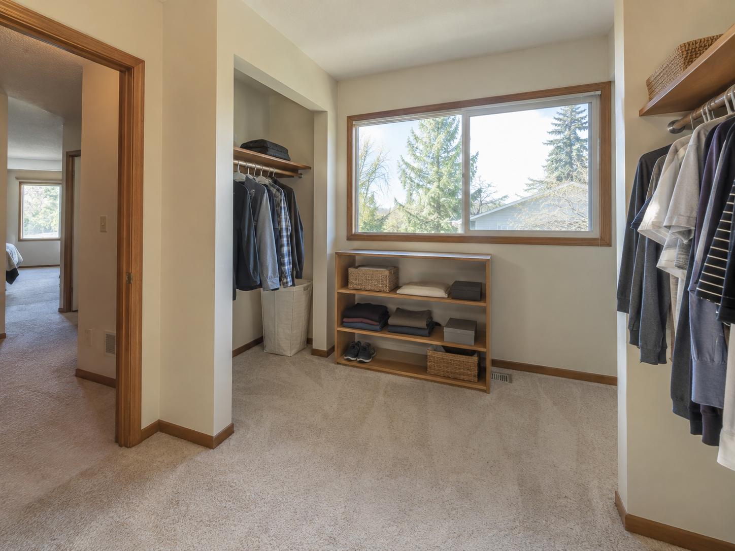 Modest walk-in closet with beige carpet, wood trim, hanging clothes on rods and a freestanding three-shelf unit under a large window.