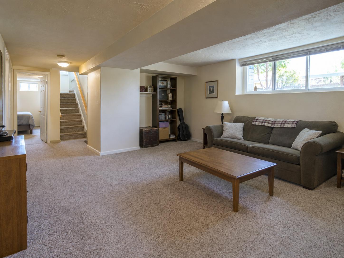 Carpeted basement with dark three-seat sofa, plain wood coffee table, open shelf niche with boxes and guitar, small high window