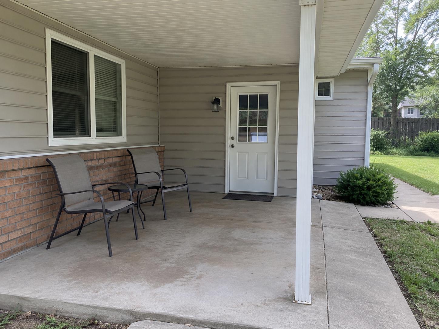 Covered patio with concrete floor, two metal mesh chairs, small side table, beige siding, white door and support column.