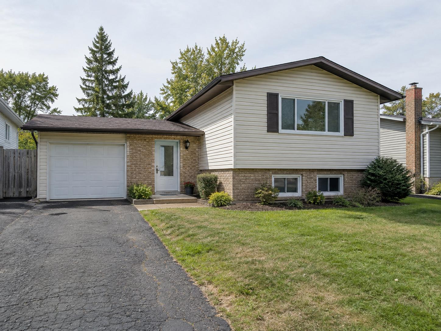 Modest split-level front exterior with light vinyl siding, brick lower wall, single-car garage, plain concrete step and sparse shrubs.