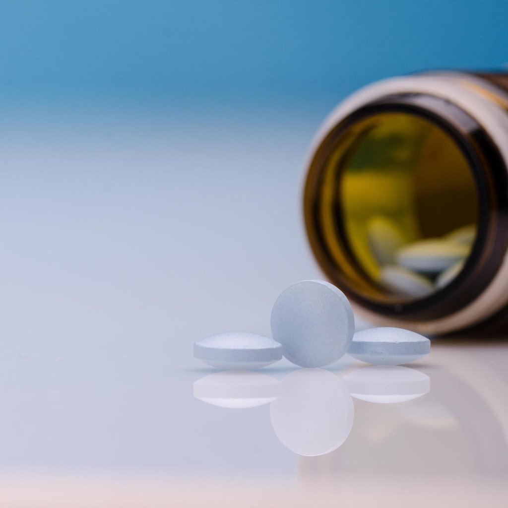 Drei blaue Viagra Tabletten liegen auf einer reflektierenden Oberfläche, während eine braune Pillenflasche im Hintergrund liegt. Die Flasche ist geöffnet, und weitere Tabletten sind darin sichtbar.