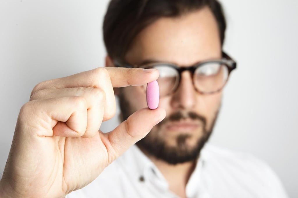 Ein Mann mit Brille und Bart hält eine rosa Viagra-Pille zwischen Daumen und Zeigefinger. Sein Gesicht ist leicht unscharf, während der Fokus auf der Pille liegt.